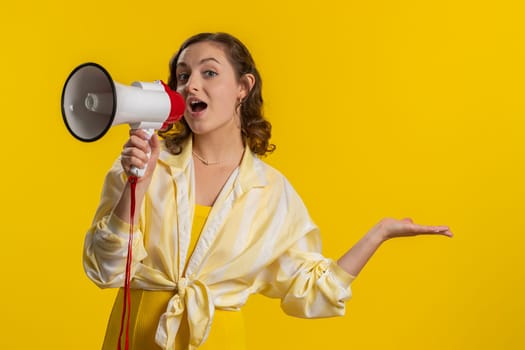 Woman talking with megaphone, proclaiming news, loudly announcing advertisement, warning using loudspeaker to shout speech, pointing empty place. Copy-space. Pretty brunette girl on yellow background