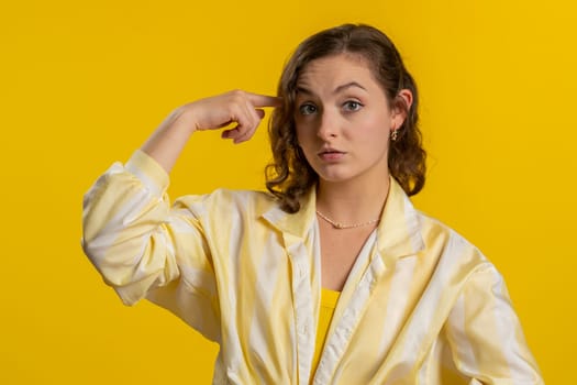 You are crazy, out of mind. Displeased woman pointing at camera, show stupid gesture finger near head, blaming some idiot for dumb insane plan idea, senseless talk. Girl isolated on yellow background