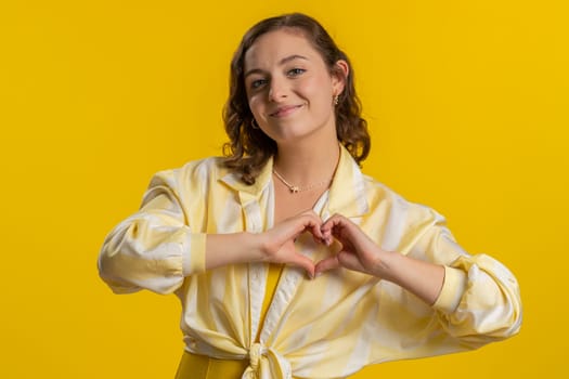 Woman in love. Smiling attractive woman makes heart gesture demonstrates love sign expresses good positive feelings and sympathy. Pretty brunette young girl isolated on yellow studio wall background