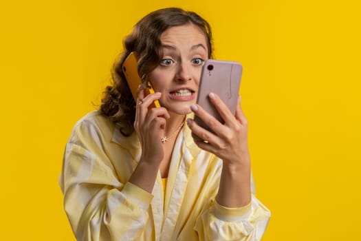 Irritated nervous stressed young woman talking screaming on two mobile phones having conversation conflict quarrel complaint dispute discuss solve problem. Girl isolated on yellow studio background