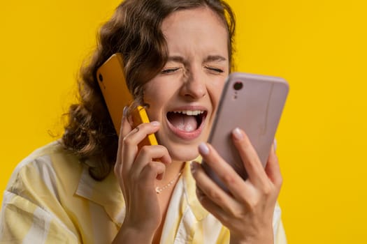 Irritated nervous stressed young woman talking screaming on two mobile phones having conversation conflict quarrel complaint dispute discuss solve problem. Girl isolated on yellow studio background