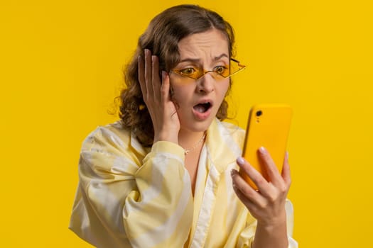 Woman use mobile phone smartphone typing browsing, loses becoming surprised sudden lottery results, bad fortune, loss, fail. Pretty brunette girl isolated alone on yellow studio background, indoors