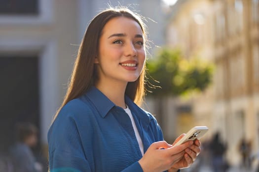 Portrait of young woman using smartphone typing text messages looking for a way on map in mobile navigator app outdoors, social media app scroll. Girl walking in urban city sunny street. Lifestyles