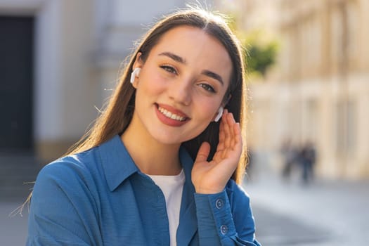 Happy relaxed overjoyed young pretty woman in wireless earphones choosing listening favorite energetic disco rock n roll music dancing looking at camera outdoors. Tourist girl walking in city street