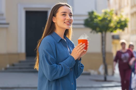 Portrait of pretty young woman enjoying morning aroma coffee steam or tea hot drink outdoors. Relaxing, taking a break. Lovely girl walking in urban city sunny street sun rays, drinking coffee to go