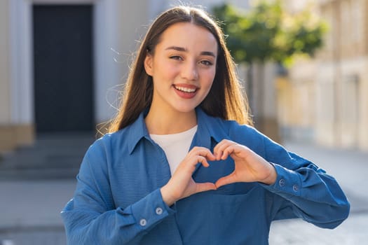 I love you. Yowoman makes symbol of love, showing heart sign to camera, express romantic feelings, express sincere positive feelings. Charity, gratitude, donation. Outdoors in sunny city street