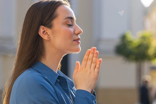 Portrait of young woman praying with closed eyes to God asking for blessing, help, forgiveness outdoors. Pretty girl clasping hands wishing luck in sunny urban city street. Town religion lifestyles