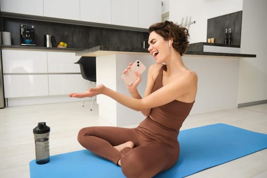 Carefree fitness girl, workout instructor sits at home with water bottle, yoga mat, wears tracksuit, sings into her smartphone, enjoys music in wireless headphones, listens song during pilates yoga.