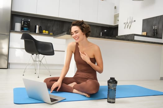 Sport and women. Young fitness instructor, yoga girl sits on rubber mat in her kitchen, waves hand and says hello at laptop, has online video gym class from home.