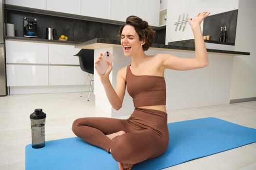 Workout from home. Happy young brunette fitness girl, sits on kitchen floor, yoga mat, with water bottle, sings while does workout exercises in wireless headphones.