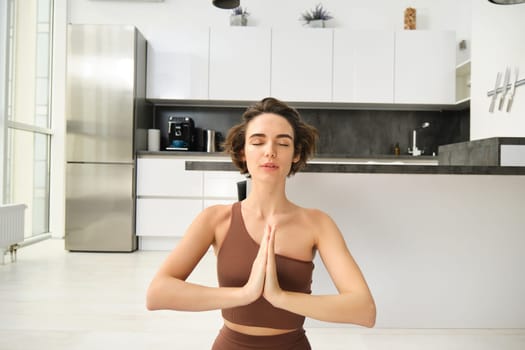 Close up portrait of young woman at home, sitting and meditating, practice yoga, making lotus pose, exercise indoors. Sport and women wellbeing concept