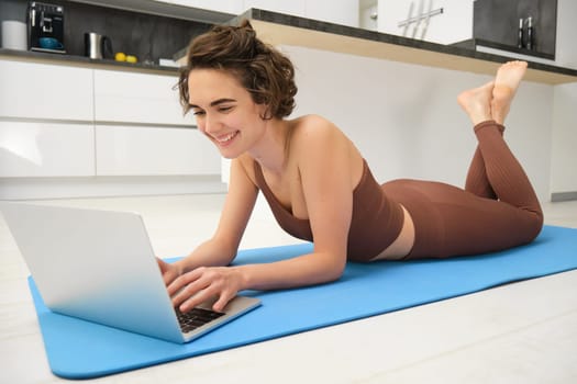 Portrait of woman athlete, girl workout at home, watches fitness videos on laptop, lying on rubber mat and doing indoor yoga session. Sport and wellbeing concept