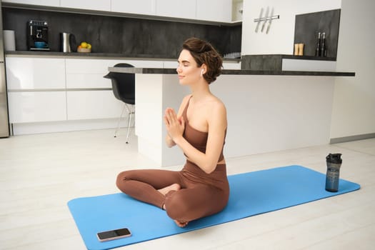 Portrait of fitness girl in kitchen, sits on yoga mat, meditates, listens meditation music in headphones, uses workout app on smartphone, does exercises at home.