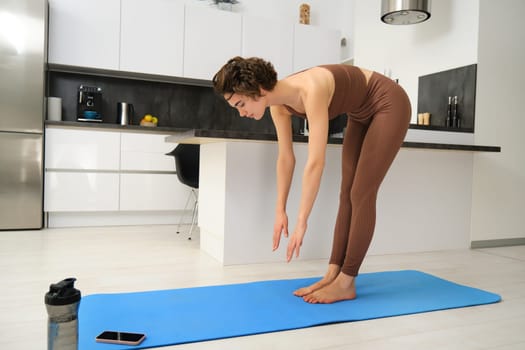 Woman follows gym video tutorial on smartphone while workout at home. Fitness girl does training at home on rubber yoga mat.