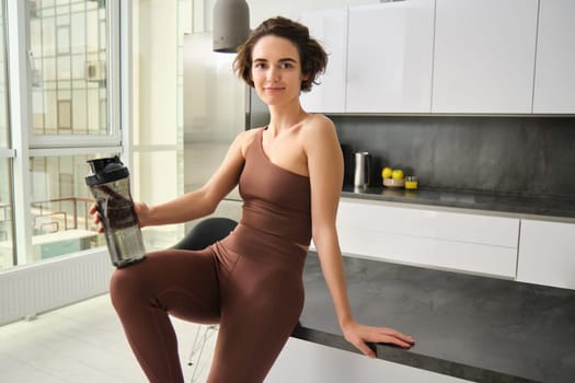 Portrait of smiling fitness girl, wearing sportswear for jogging, workout training, standing in kitchen and drinking water from sports bottle, looking fit and happy.