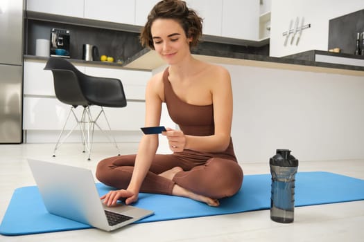 Image of young fitness girl, female athlete at home, workout on kitchen floor, holding credit card with laptop, purchasing online training session, gym application for home workout.