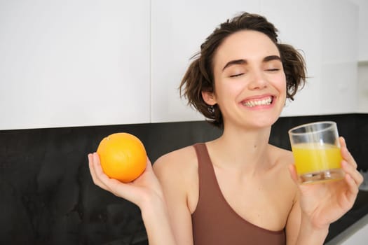 Sport and healthy lifestyle. Smiling happy young woman, holding an orange, drinking fresh juice and laughing, wearing activewear, standing in kitchen. Copy space