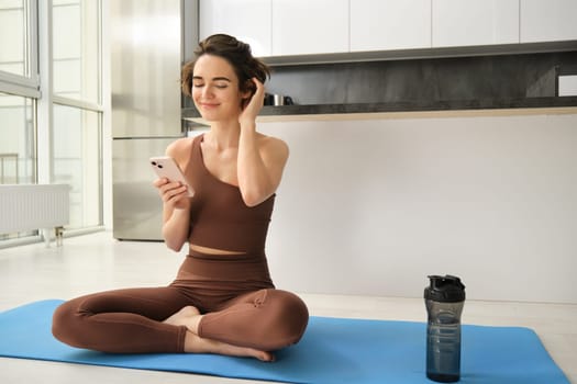 Sport and women lifestyle. Young woman does workout at home, sits on yoga mat with smartphone in bright kitchen, wears activewear, uses mobile phone app for indoor exercises.