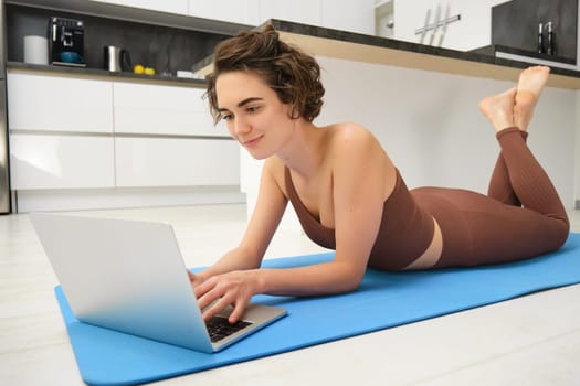 Portrait of beautiful sportswoman, fitness girl watching videos on laptop during workout, follow online pilates videos while exercise at home, wearing activewear in kitchen.