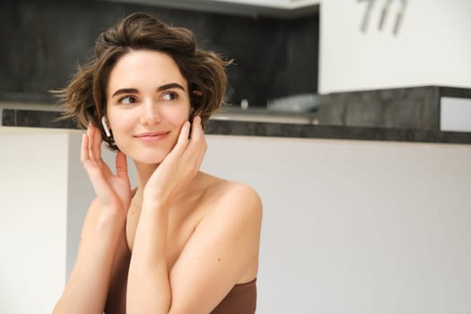 Close up of young smiling brunette woman, fitness girl in kitchen, looks aside, touches her face, listens to mindful app training session in wireless earphones, workout at home.