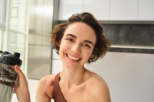 Lifestyle, sport and health concept. Portrait of happy beautiful woman, holding water bottle and smiling, wearing workout clothes at home, doing yoga exercises indoors.