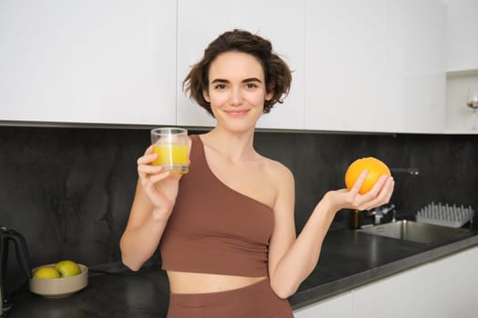 Healthy lifestyle and sport. Beautiful smiling woman, drinking fresh orange juice and holding fruit in her hand, enjoying her vitamin drink after workout at home, standing in kitchen.