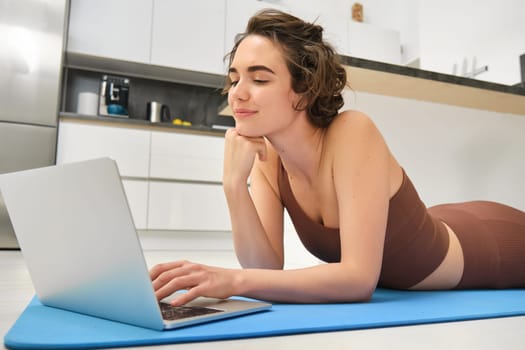 Portrait of beautiful sportswoman, fitness girl watching videos on laptop during workout, follow online pilates videos while exercise at home, wearing activewear in kitchen.