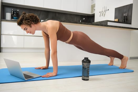 Portrait of young sportswoman in activewear, standing in plunk, following online video gym class on laptop, drinking water during her workout training session at home, exercising in kitchen.