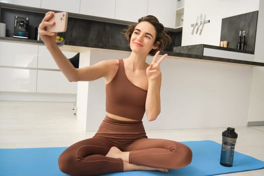 Portrait of young fitness woman, sitting at home on rubber mat, records video on smartphone, doing workout vlog, making live online training session from home. Copy space