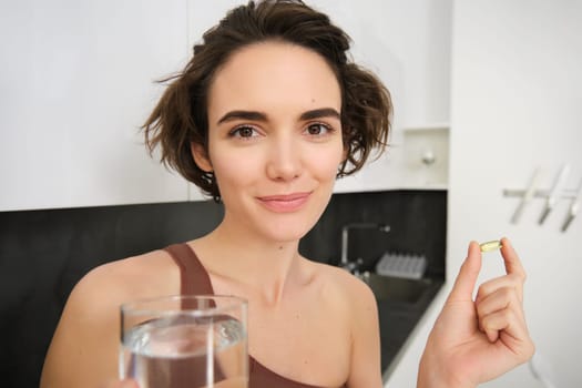 Portrait of sportswoman drinking water, taking vitamins, dietry supplements for healthy skin, having omega-3 pill, standing in her kitchen in workout clothing.