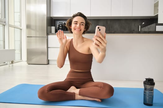 Smiling fitness girl, young athlete sits at home on yoga mat, waves hand at smartphone, joing online workout training session from home, says hello.