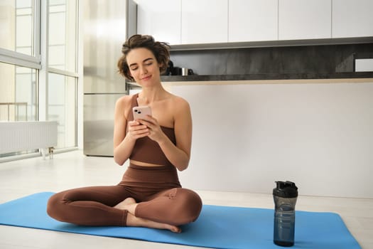 Yoga and sport at home concept. Young woman sitting in bright rome with mobile phone, doing workout on rubber mat, searching for exercises online on smartphone app, using sports equipment.