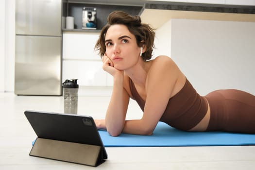 Thinking girl in sportswear, does fitness training at home, lies on rubber mat with tablet, watches workout video tutorials and looking complicated.