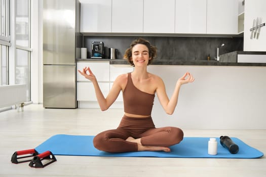 Smiling girl athlete, sportswoman doing yoga at home in activewear, sitting on yoga rubber mat in lotus pose, meditating, practice mindfulness exercises.