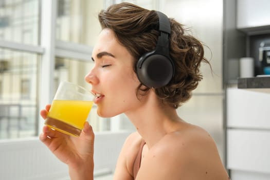 Smiling sportswoman, fitness girl does workout at home, wears headphones, listens music and drinks orange juice after training session.