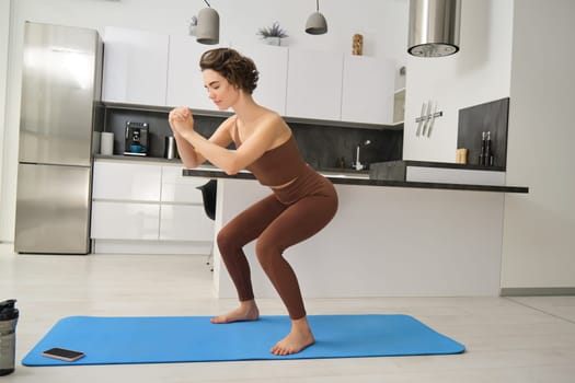 Image of young woman doing squats at home, workout on rubber mat in bright room indoors, wearing activewear for training. Copy space