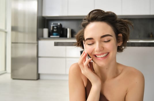 Close up portrait of happy smiling woman talking on mobile phone from her kitchen. Chatty girl with smartphone, laughing, calling someone. Lifestyle and women concept