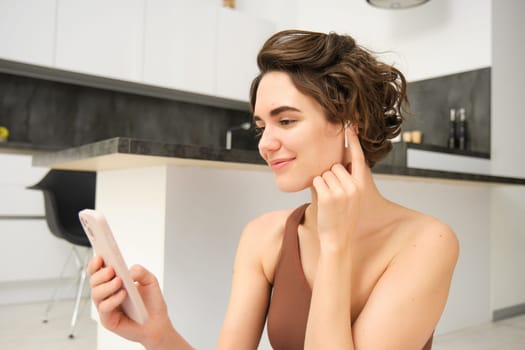 Close up of young fitness instructor, healthy and fit woman with smartphone, wireless headphones, records her workout training session at home, connects to client on video call for yoga classes.