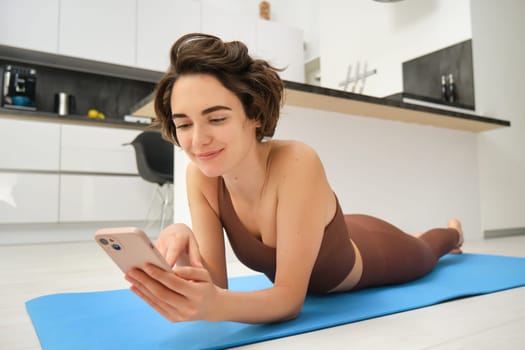 Portrait of fitness woman lying on yoga mat, looking at smartphone, using mobile phone during workout.