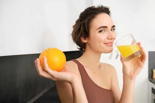 Image of sportswoman, fitness girl holding glass of juice and an orange, smiling, drinking vitamin beverage after workout, standing in her kitchen at home. Healthy lifestyle and sport concept.