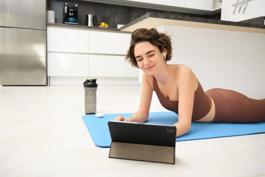 Young fitness woman, girl in tracksuit lying on fitness mat, looking at yoga, pilates video tutorial on tablet, does workout from home, uses gym app, training at home.