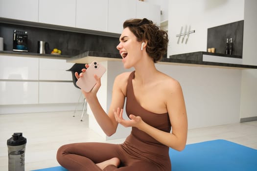 Workout from home. Happy young brunette fitness girl, sits on kitchen floor, yoga mat, with water bottle, sings while does workout exercises in wireless headphones.