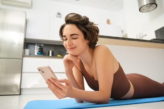 Image of sporty girl resting on yoga rubber mat, workout at home, using her smartphone, looking for online fitness tutorials, mobile app for training.