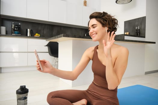 Young fitness girl at home, sport instructor in tracksuit waves hand at smartphone, sits on workout yoga mat, waves hand at mobile camera, says hi to online gym class, exercise remotely.
