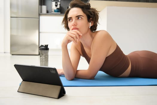Portrait of fitness girl with thoughtful face, lying on rubber yoga mat at home, listening and watching sport workout video tutorial on tablet, thinking.