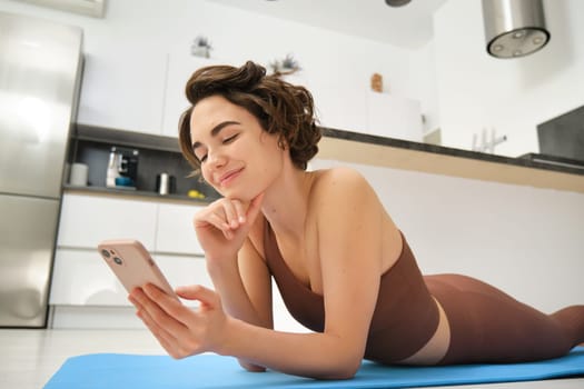 Online fitness and wellbeing. Portrait of young woman lying on yoga mat, looking at smartphone app, looking at workout tips, doing training at home in activewear.