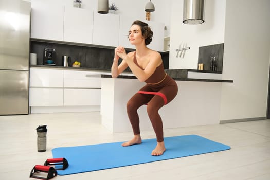 Fitness at home. Young sportswoman, girl in activewear, doing squats in her kitchen, workout indoors on rubber mat, using elastic rubber band for exercises.