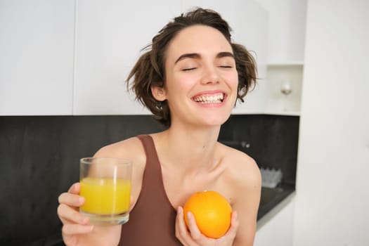 Healthy lifestyle and sport. Beautiful smiling woman, drinking fresh orange juice and holding fruit in her hand, enjoying her vitamin drink after workout at home, standing in kitchen.