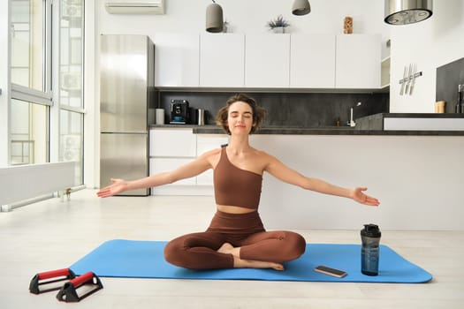Mindfulness and meditation. Young relaxed fitness woman practice yoga at home, sitting in meditation pose, lifting hands up to the air and doing breathing exercises on rubber mat.