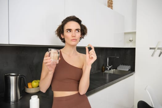 Portrait of sportswoman drinking water, taking vitamins, dietary supplements for healthy skin, having omega-3 pill, standing in her kitchen in workout clothing.